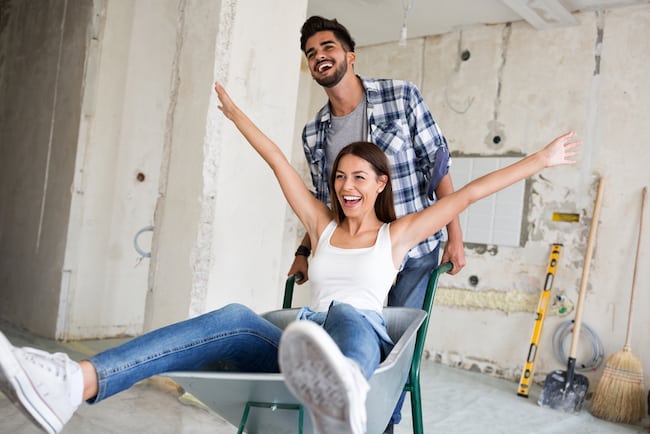 A young couple plays around during a bathroom remodel. The women sits in the wheelbarrow and the man pushes her.